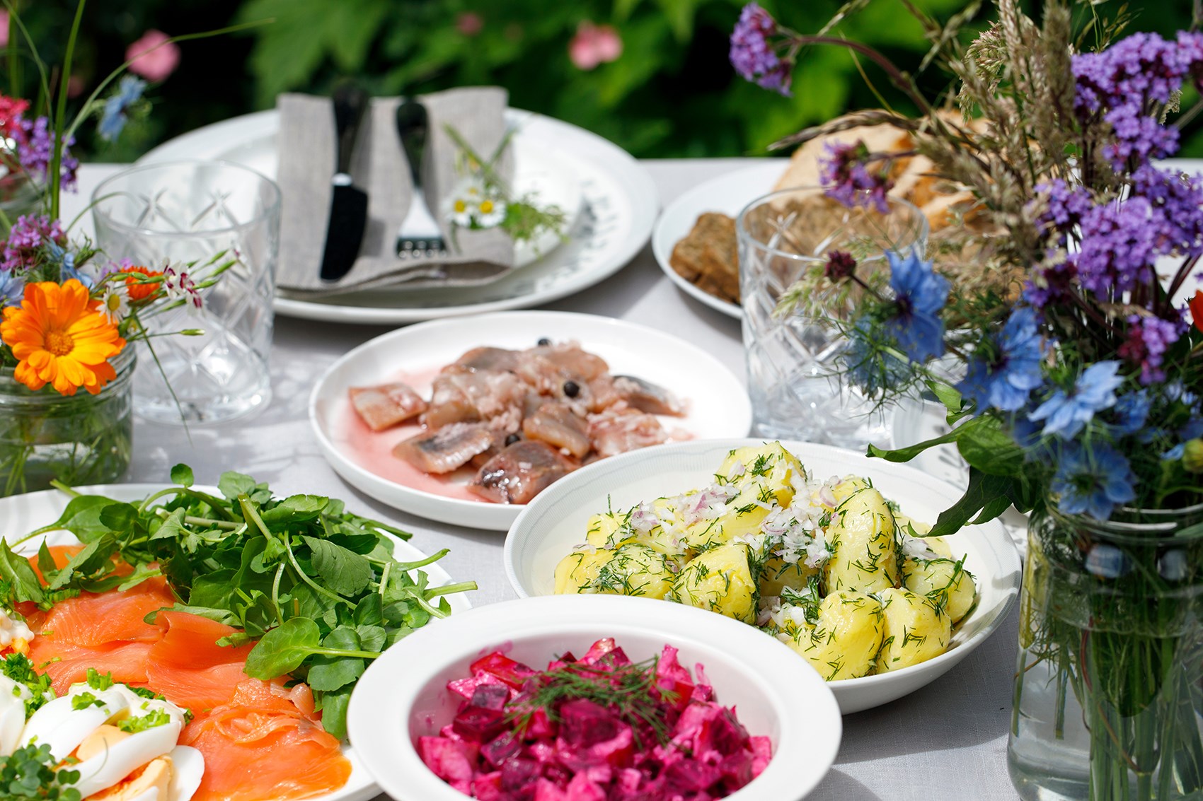Olika tallrikar dukat på ett bord med sill, potatis och lax samt blommor runtom. 
